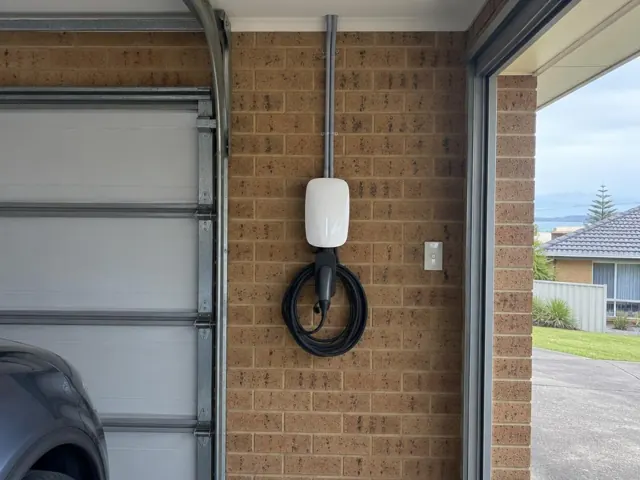 Wall-mounted EV charger on a brick garage wall beside a roller door in a suburban home in Long Jetty